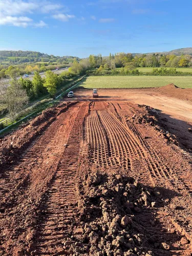 A freshly leveled, reddish-brown dirt road under construction, surrounded by green fields and trees. A small construction vehicle is visible in the distance, with a blue sky and hills in the background.