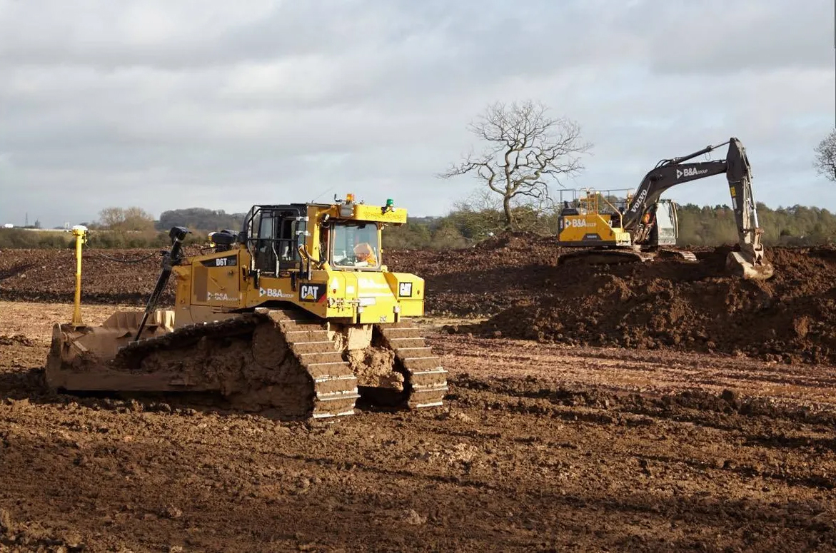 A yellow bulldozer in the foreground levels a muddy construction site, while an excavator in the background moves soil; a bare tree and cloudy sky are visible in the distance.
