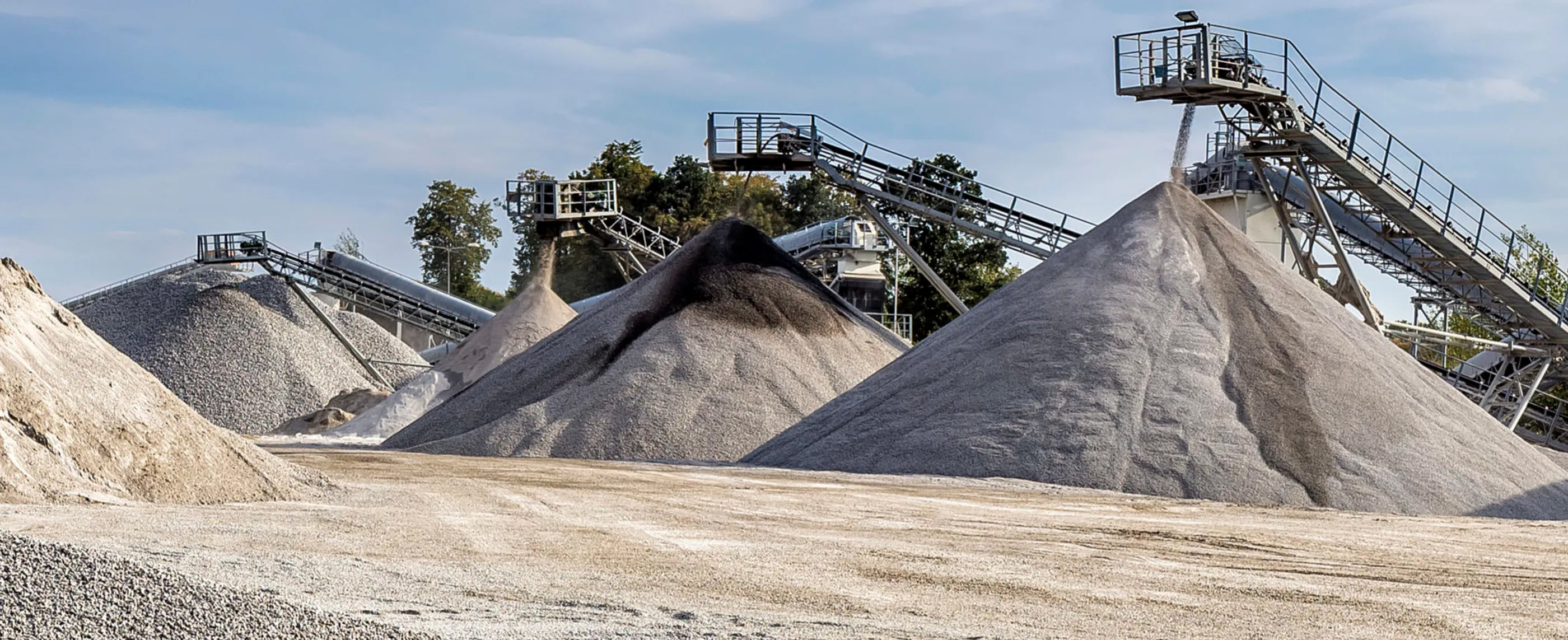 Piles of gravel and sand of varying sizes are arranged in an open quarry, with conveyor belts and machinery in the background against a blue sky.