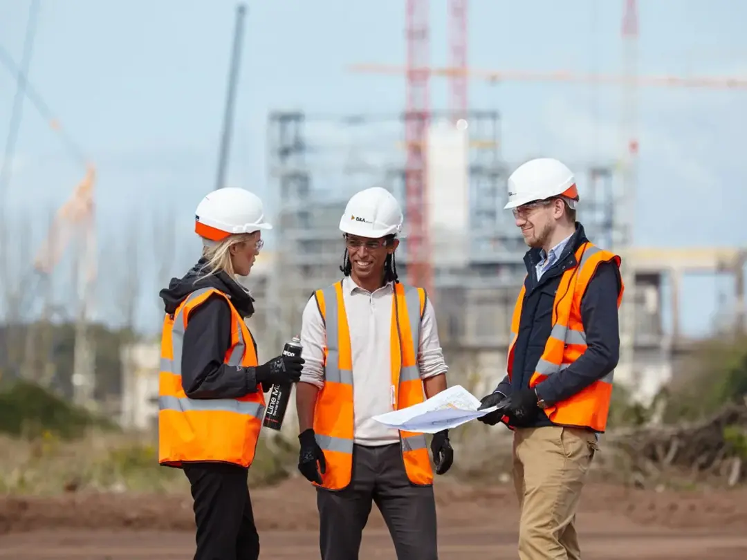Three construction workers wearing safety helmets and orange vests stand on an outdoor job site, discussing a blueprint. A partially constructed building and cranes are visible in the background.